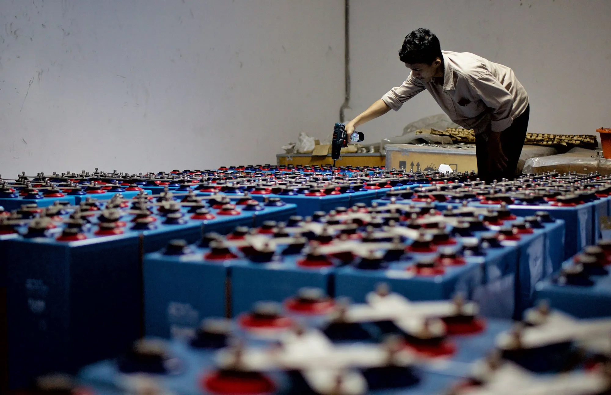 Rows of blue battery cases on a factory floor, a worker filling cells at the far end.