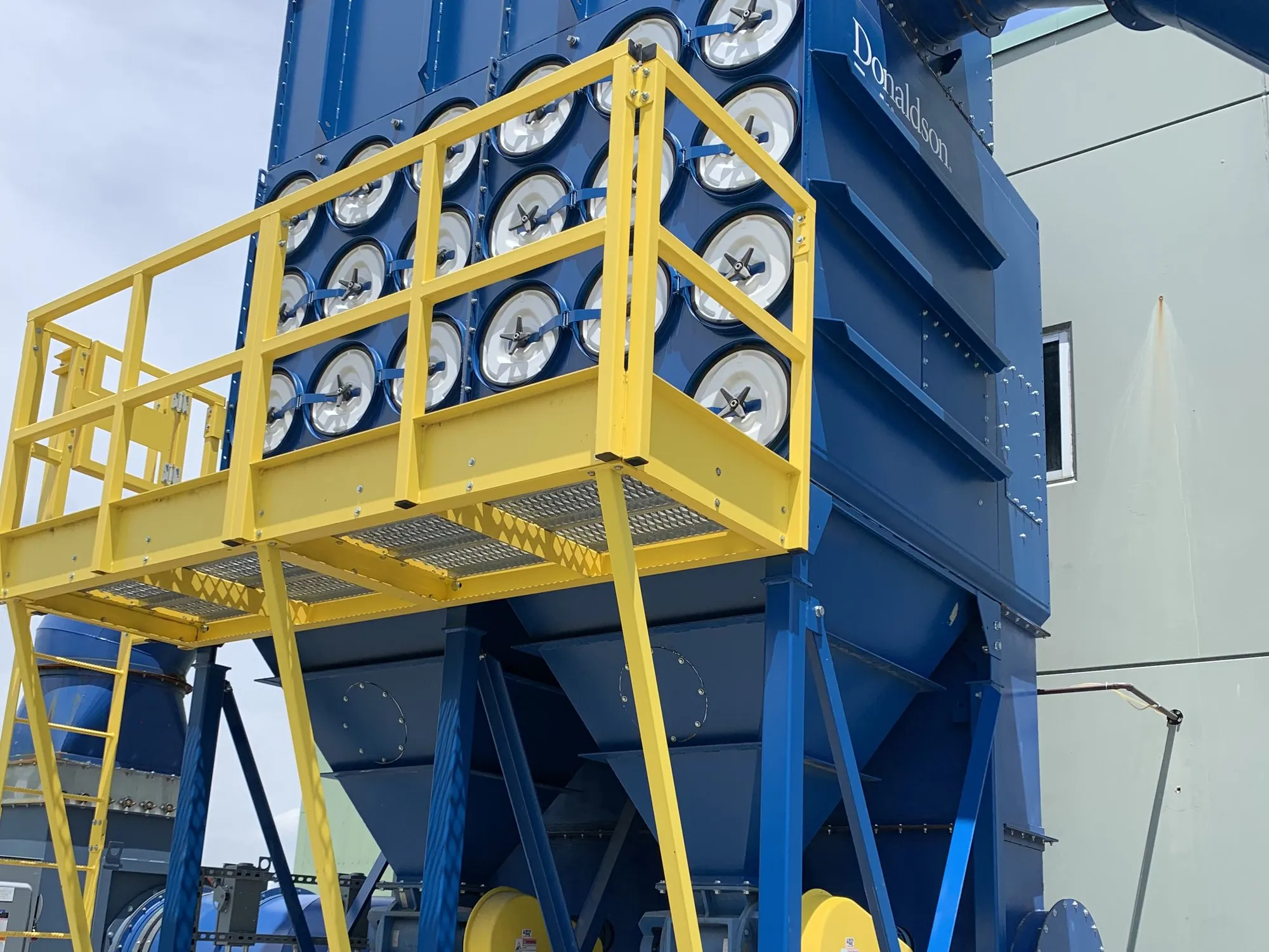 A blue Donaldson cartridge collector outdoors on structural steel, with a yellow access catwalk and ladder.