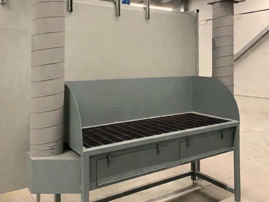 A grey downdraft bench with a mesh work surface and back plenum, spiral ducting rising behind it to the roof.