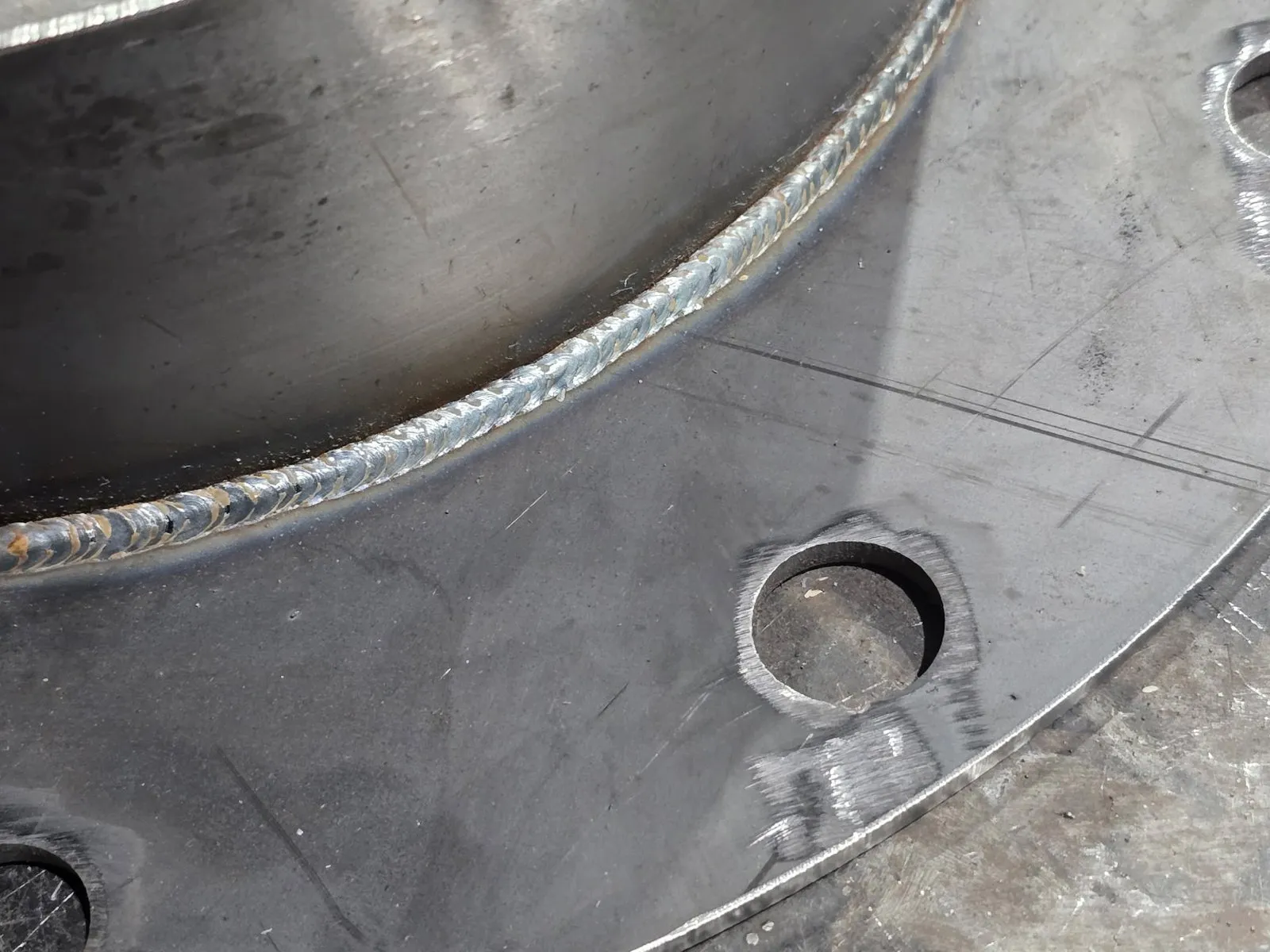A welded seam running around the flange of a large rolled duct section on the shop floor, bolt holes punched through the plate.