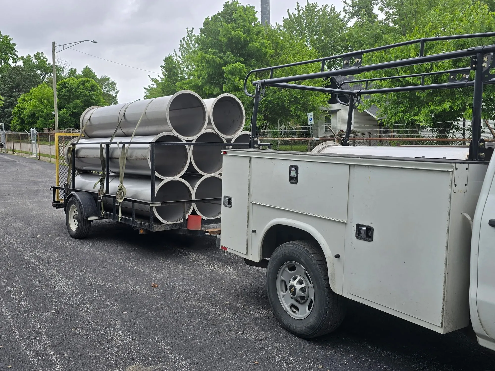 Lengths of large-diameter spiral duct strapped to a trailer behind a white service truck, ready to leave the shop.