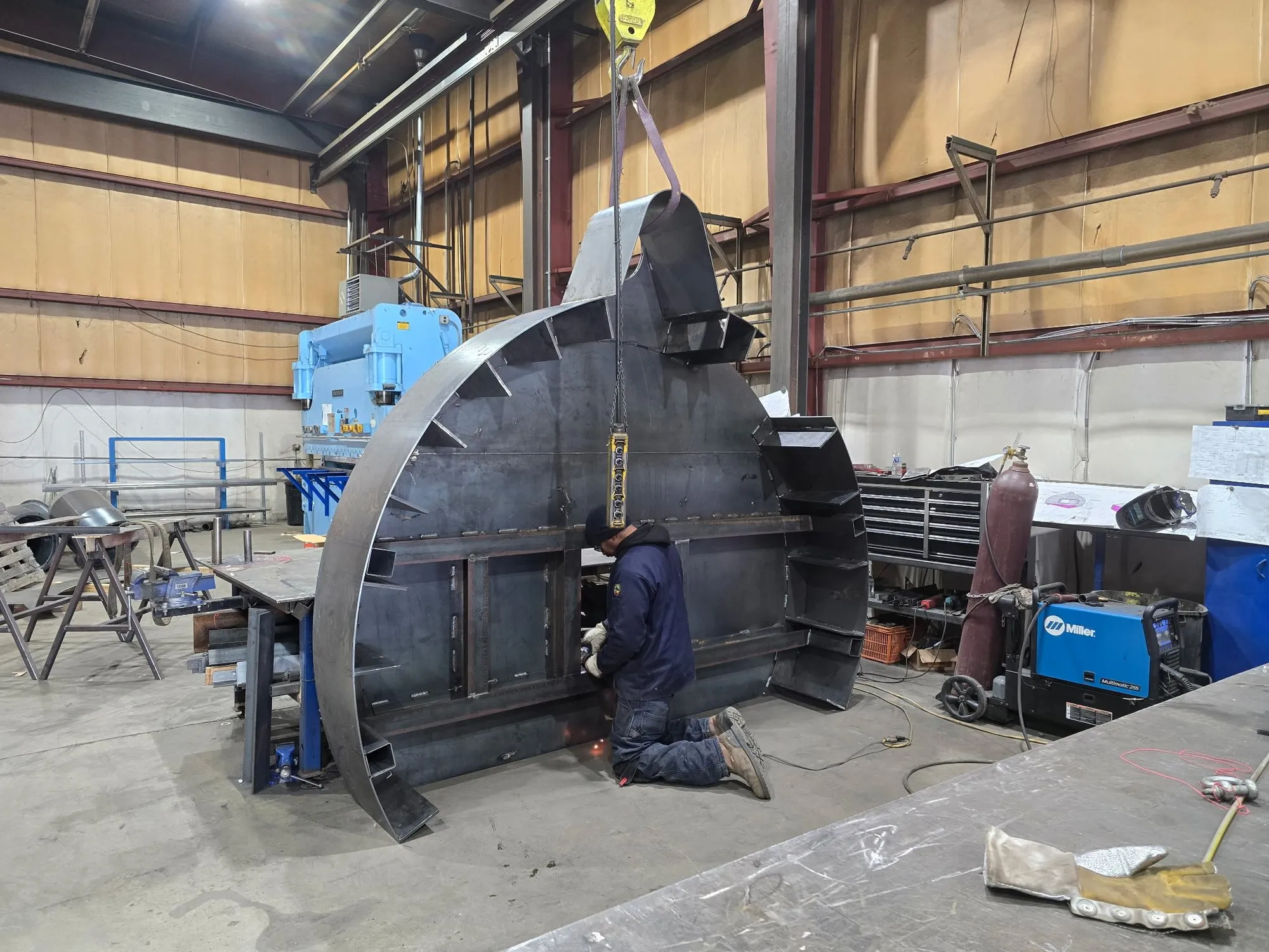 A DCG fabricator kneeling to weld the internal framing of a large round furnace lid, slung from the shop crane.