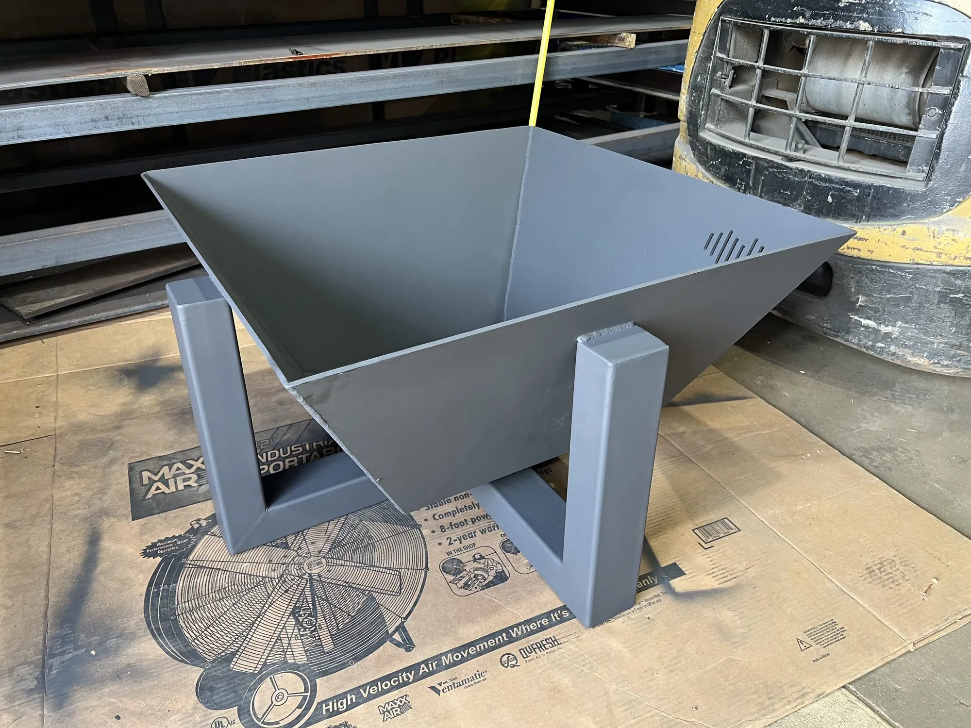A fabricated steel fire pit with a folded square bowl on welded angle legs, painted grey, on the shop floor.