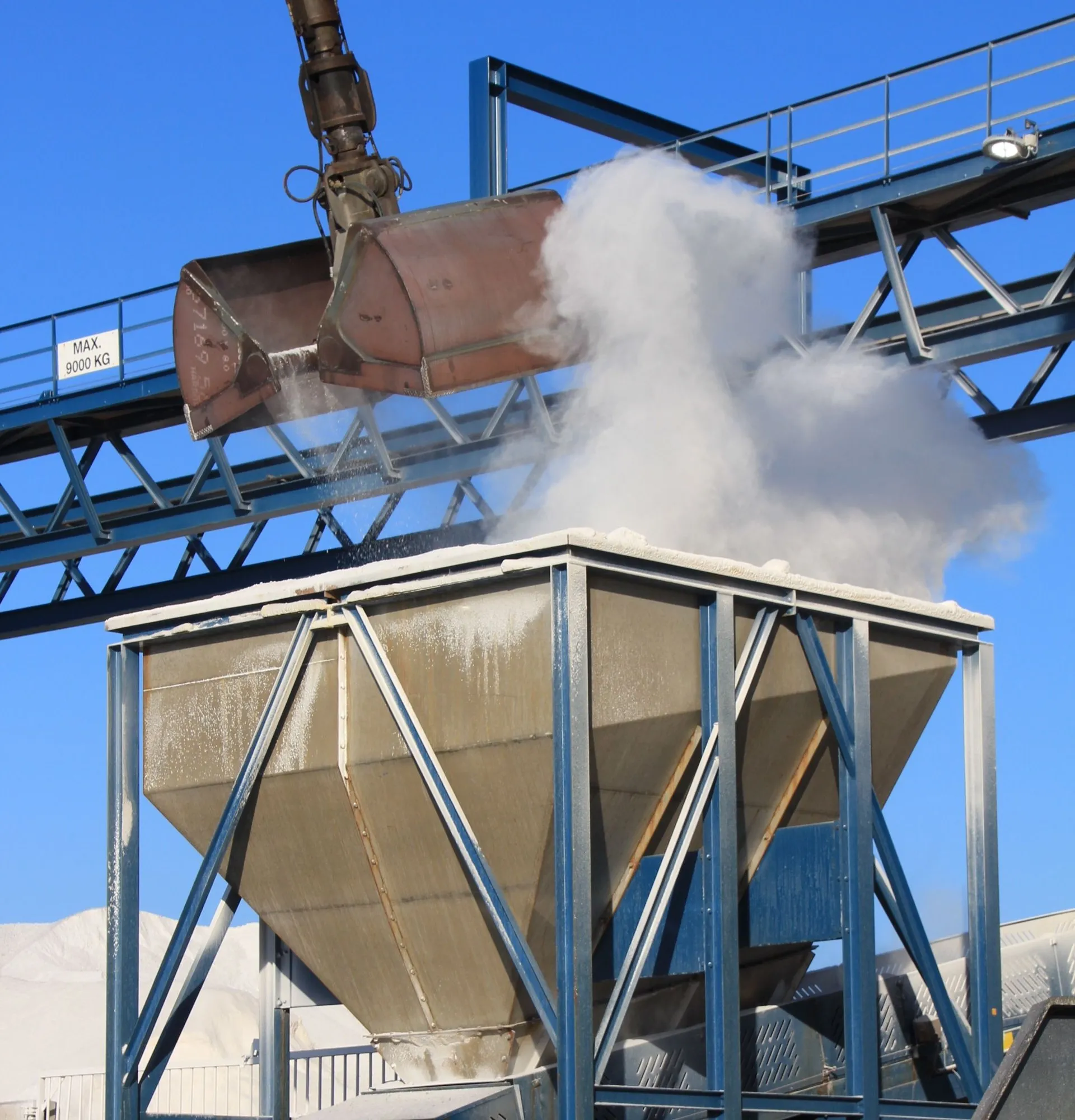 A grab bucket discharging bulk powder into a steel hopper, a cloud of dust rising off the transfer.