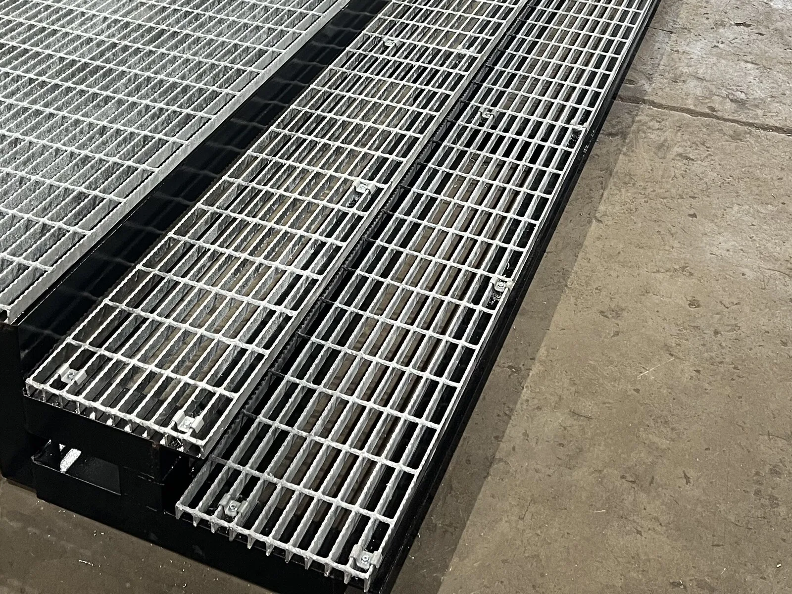 Close-up of galvanized grating panels clipped onto black welded steel frames, laid out on the shop floor.