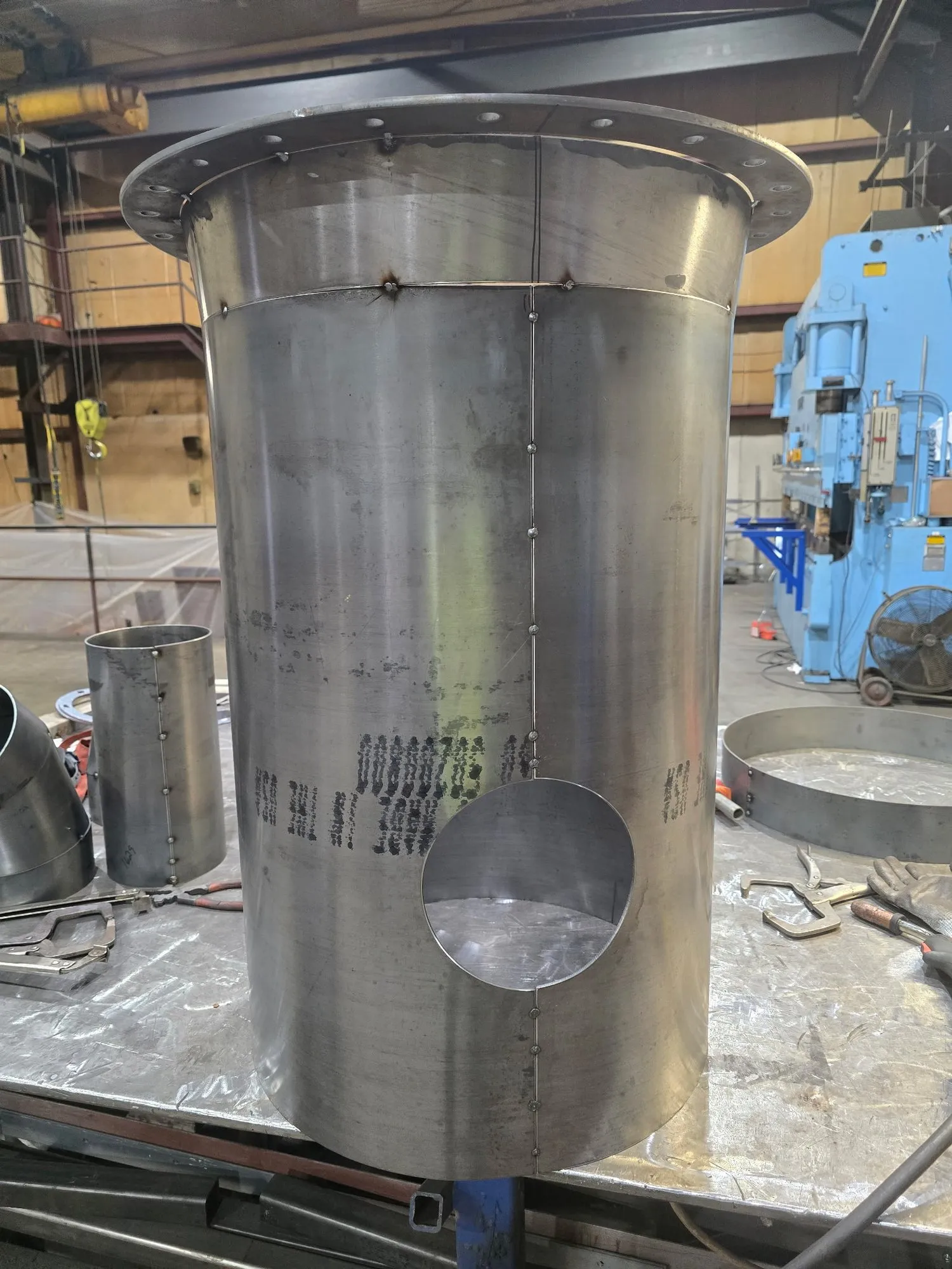 A rolled stainless shell with a bolted top flange and a cut branch opening, standing on the shop floor beside a press brake.