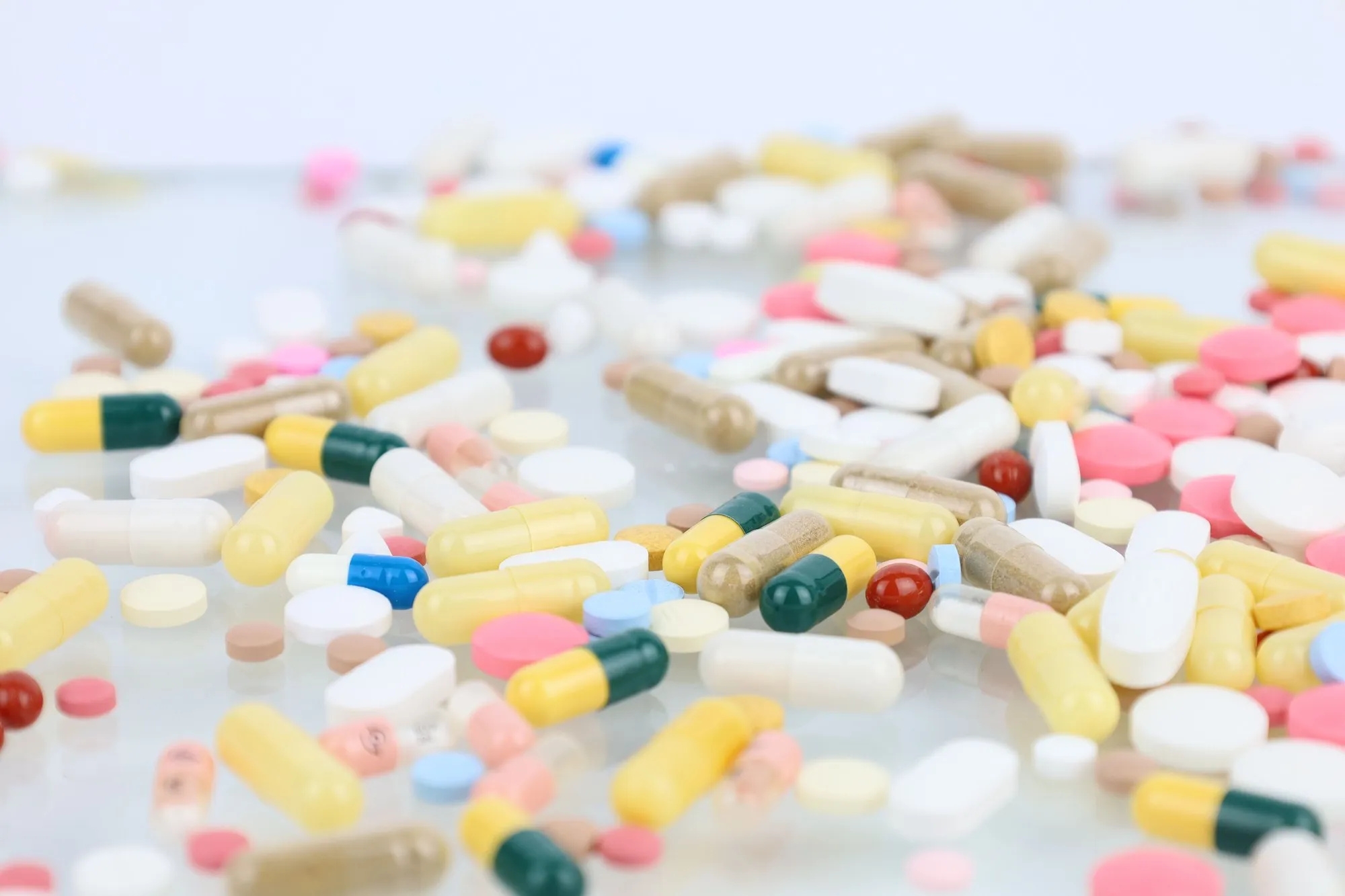 Loose pharmaceutical tablets and capsules in many colours scattered across a white surface.