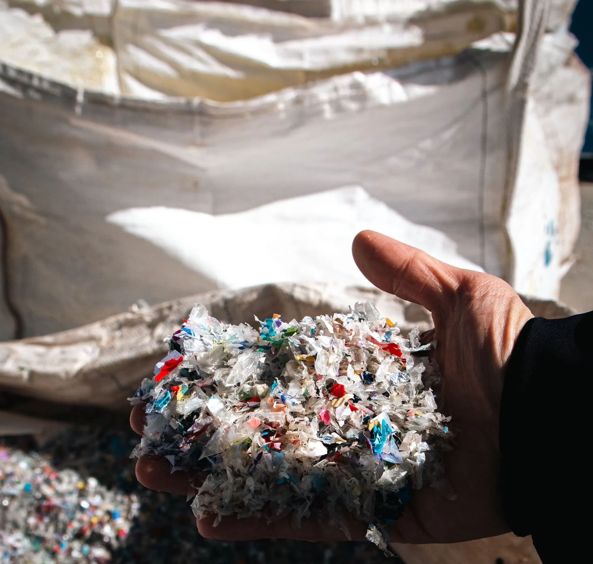 A hand holding a heap of shredded mixed-colour plastic regrind in front of a large white bulk bag of the same material.