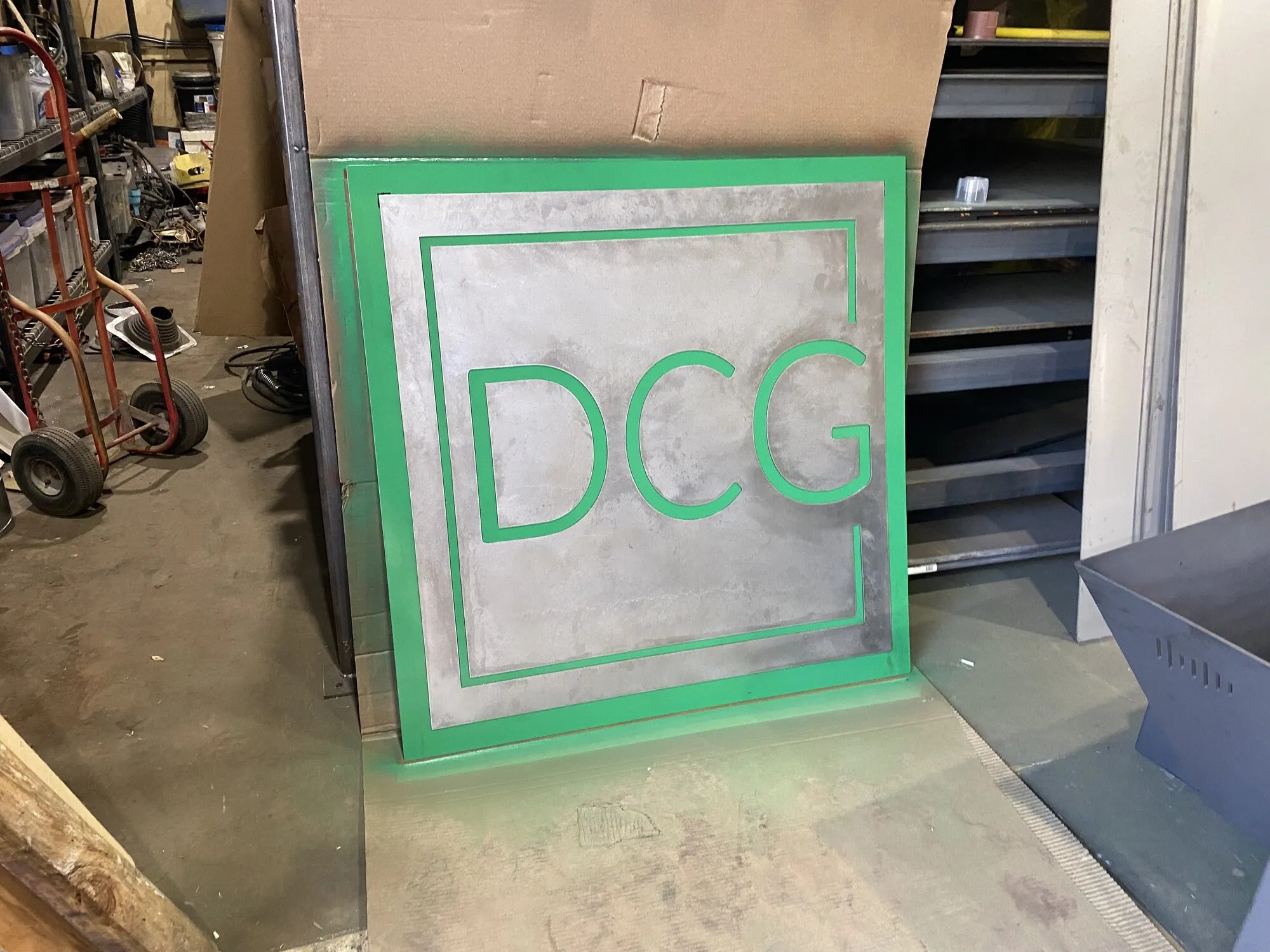 A square steel panel with the DCG logo cut through it, its frame and letters painted green, leaning against a wall in the shop.