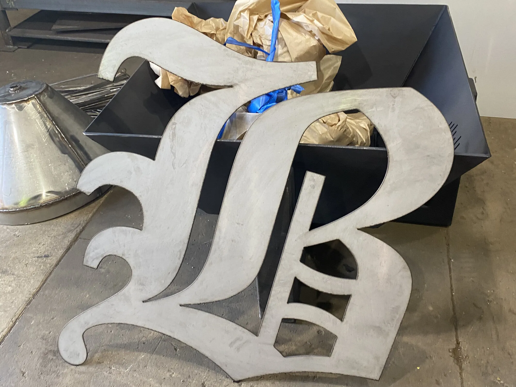 A large blackletter monogram cut from steel plate, propped against a fabricated steel bowl on the shop floor.
