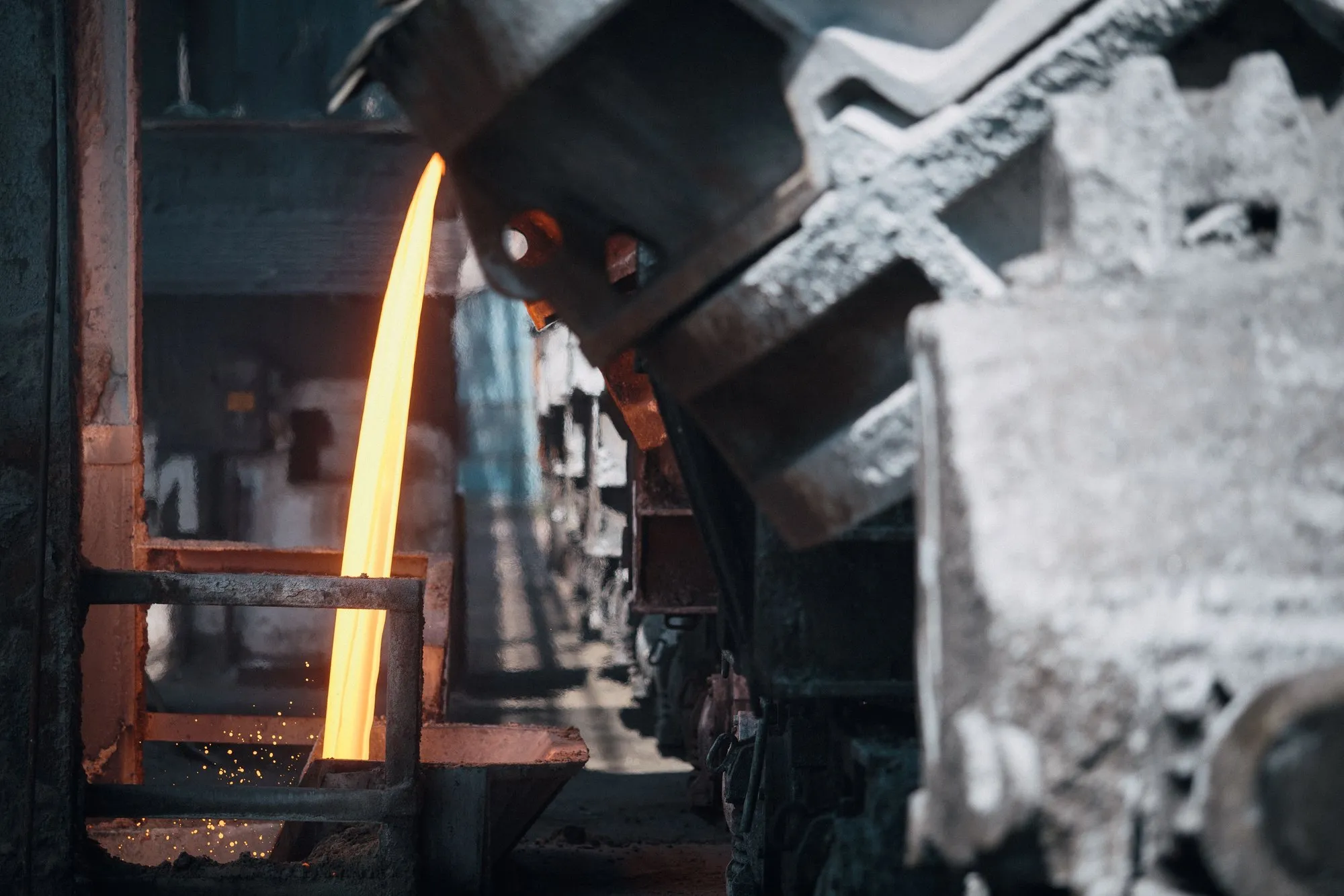 Molten metal pouring in a bright stream from a tilted furnace ladle on a foundry floor.