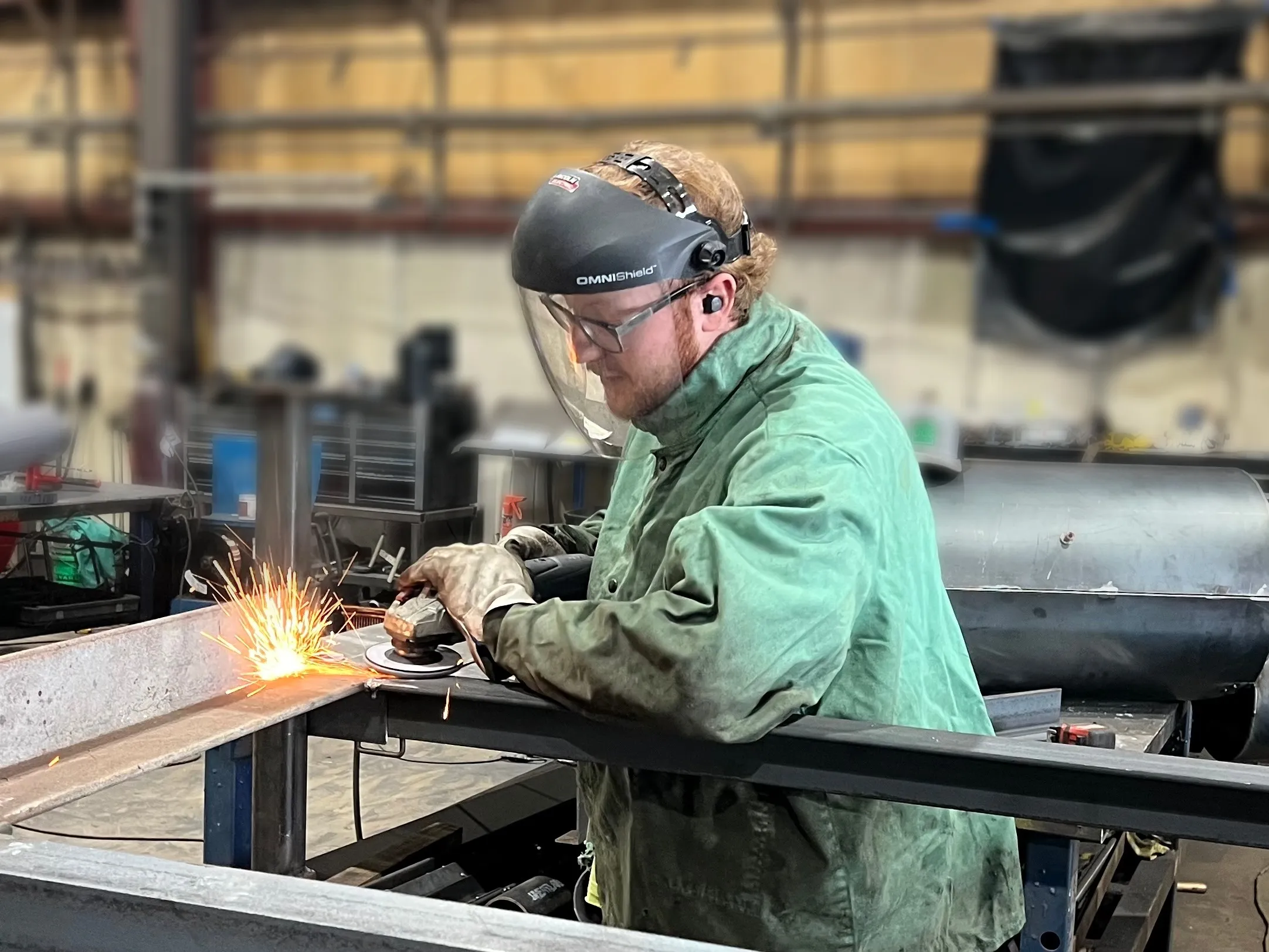 A DCG fabricator in a welding jacket and face shield grinding a steel frame, sparks flying.