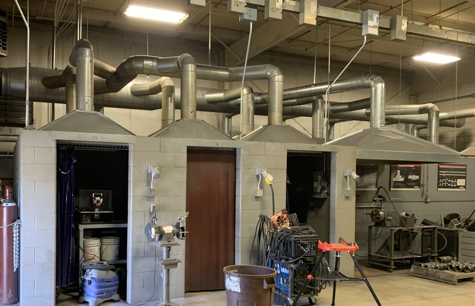 A row of block-built welding booths with fabricated capture hoods, spiral ducting running overhead to collect from each one.